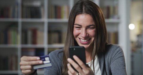 Cinematic shot of young happy smiling woman is entering credit card information by using smart phone while is making shopping online in living room at home.