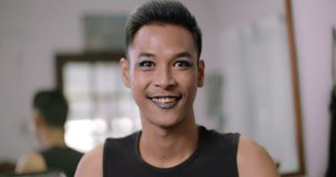 A smiling short-haired Asian gay who owns a small beauty salon in the village wearing false eyelashes and make up his face.