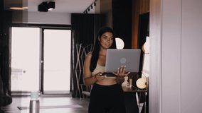 A young dark-skinned brunette with a laptop in her hands walks through the kitchen and talks video communication, participates in a video conference, undergoes online training. Covid-19. Quarantine.
