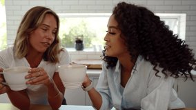 Mixed race female friends chatting together smiling enjoying hot coffee in kitchen. 
