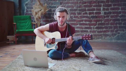 Man acoustic guitar sitting floor with laptop and headphones playing singing song online music lessons. Caucasian young guitar player practicing musical instrument video call Serenade for girlfriend