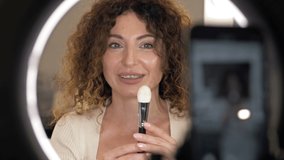 Beautiful, middle-aged beauty blogger talks about the types of makeup brushes and their purpose. Woman sitting in front of a mirror in which a video camera is reflected.