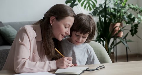 Homeschooling writing and drawing a little cute boy pupil in a bright apartment. Young attractive mother teacher helping smart preschooler son learn to count and develop his math skills