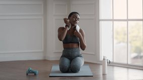 Tired Black Sporty Lady Wiping Her Sweat With Towel After Intensive Domestic Training, Exhausted African American Woman Sitting On Yoga Mat, Relaxing After Fitness Workout At Home, Slow Motion