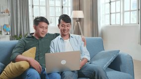 Portrait Of Asian Gentle Gay Couple Using Laptop Computer, While Sitting On A Couch In Cozy Stylish Apartment. Adult Boyfriends Online Shopping On Internet, Watching Funny Videos On Streaming Service
