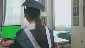 Excited Asian Woman Showing Off A University Certificate To Laptop Computer With Green Mock-Up Screen During An Online Video Call. Pretty Female Graduate Wearing A Graduation Gown And Cap Sitting On T