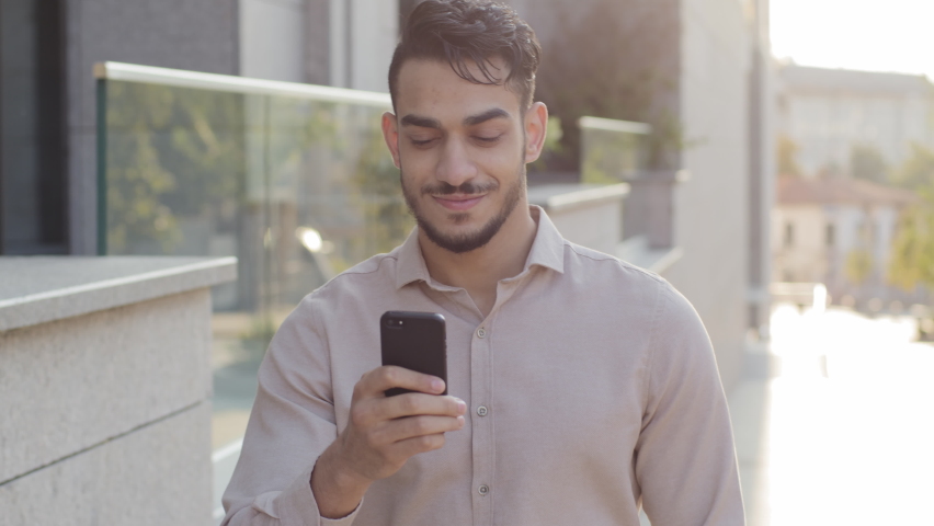 Hispanic business man smiling walking city street looking at mobile phone online map application app looking around enjoying urban view. Happy arabian guy tourist with smartphone goes outside outdoors | Shutterstock HD Video #1079602142