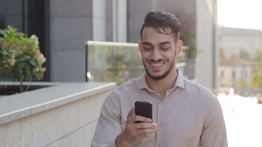 Hispanic business man smiling walking city street looking at mobile phone online map application app looking around enjoying urban view. Happy arabian guy tourist with smartphone goes outside outdoors Royalty-Free Stock Footage #1079602142