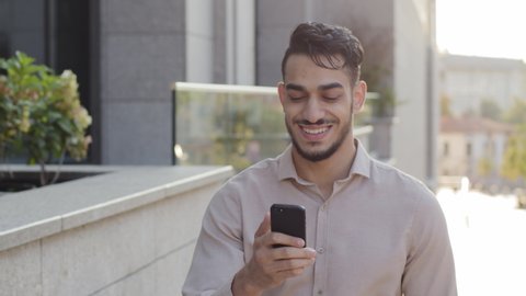 Hispanic business man smiling walking city street looking at mobile phone online map application app looking around enjoying urban view. Happy arabian guy tourist with smartphone goes outside outdoors