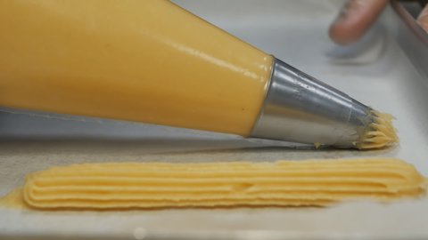Close-up, DOF, dolly shot: Squeeze the equal size custard dough for eclairs from a pastry bag into a baking tray on paper. French traditional dessert