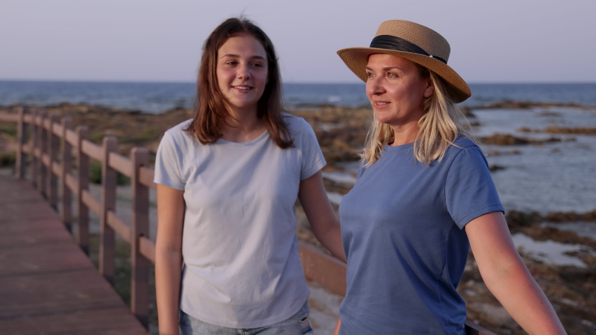 Mother and Daughter embracing on wooden sidewalk by the sea at sunset. A middle-aged woman and teenage girl standing side by side and wearing blue t-shirts and shorts. Motherhood and travel concept Royalty-Free Stock Footage #1079606795