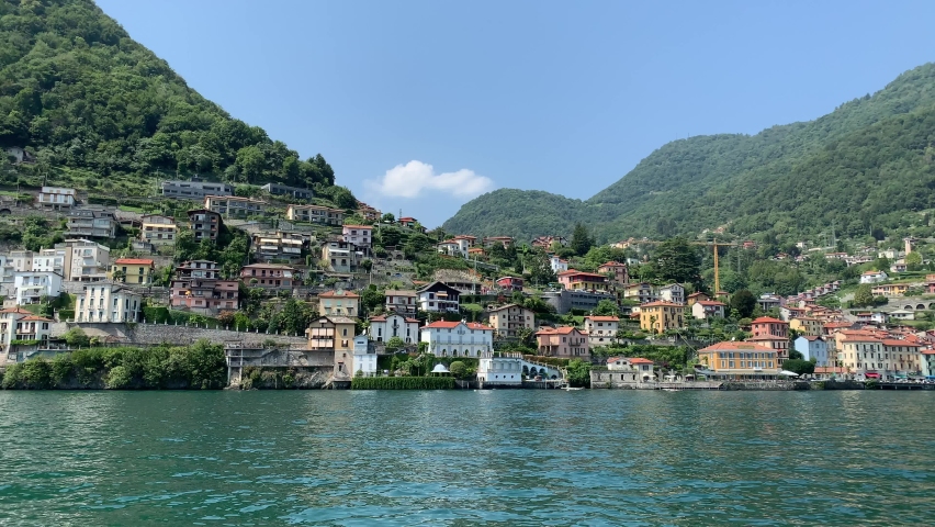 Skyline of Argegno town with houses on hills, mountains. Landscape of Como Lake (Lago di Como) shore. View from the moving boat. Motion of waves and emerald water. Argegno, Como Lake, Lombardy, Italy. | Shutterstock HD Video #1079607500