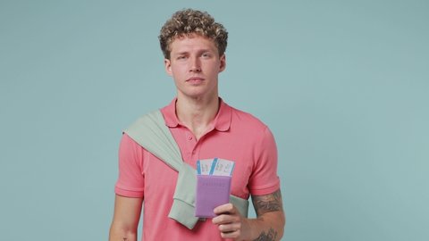 Traveler tourist young curly man 20s wear pink t-shirt hold passport boarding pass tickets isolated on plain pastel light blue background studio portrait Passenger traveling abroad on weekends concept
