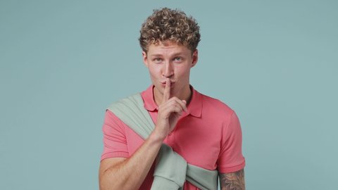 Secret attractive young curly man 20s years old wears pink t-shirt look aside say hush be quiet with finger on lips shhh gesture winking isolated on plain pastel light blue background studio portrait