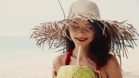 Beautiful pretty asian woman in hat and red dress drinking coconut on tropical paradise beach. Woman quench her thirst with coco nut. Beauty, baunty, travel, fashion concept. Vacation on exotic island