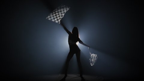 Rear view of silhouette young woman waving checkered race flag to signal the start of racing event. Brunette posing full length in dark smoky studio with backlight. Slow motion ready, 4K at 59.94fps.