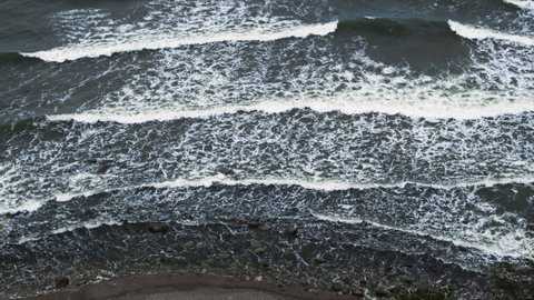 A view from above of waves crushing on a shore of the cold dark sea. Wild beach with stormy waves near cliffs and rocks in water.