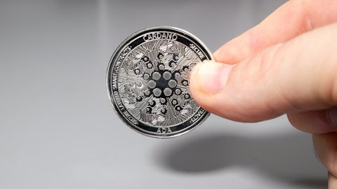 Silver cardano coin held between fingers in hand on gey table