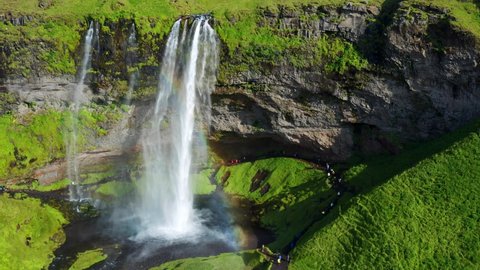 Tourists Visit Breathtaking Seljalandsfoss Waterfall With Rainbow During Summer In South Iceland. - aerial pullback