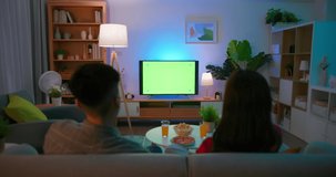 Asian young couple are watching green chroma key screen TV with food and drinks fun while sitting on couch at night in the living room