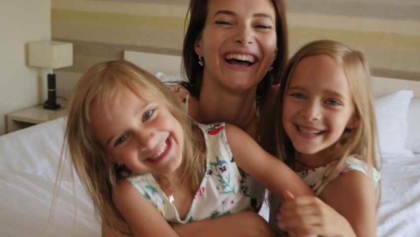 A happy mother hugs her twin daughters sitting on a bed in a hotel, with a large suitcase next to the bed. A family with twin children stay at the hotel in the summer. | Shutterstock HD Video #1079623241