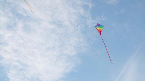 4K multicoloured kite flying in blue sky under gusts of wind with some white clouds up high. Hobbies and leisure activities concept.