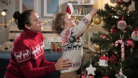 Mom and kid are decorating Christmas tree. Mother and child of 2-3 years old at home decorate Christmas tree, put toys on branches. Christmas lights are shining in background. Slow motion