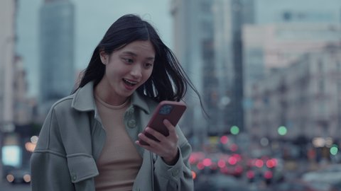 Portrait of charming young woman reading some good news on smartphone that holding in hands. Asian brunette using cell phone on street of big city.