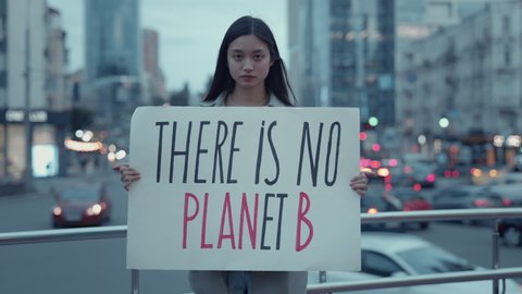 Responsible young lady with long dark hair holding banner with phrase there is no planet b. Asian woman standing on street with night light behind.