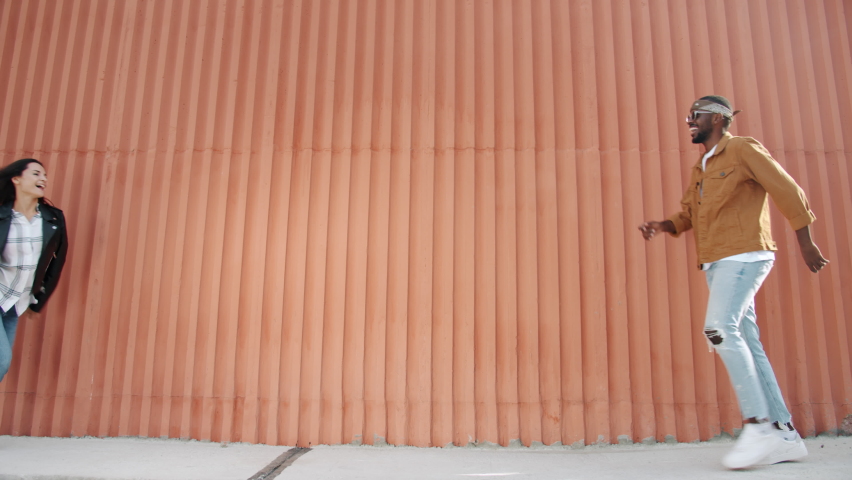 Happy mixed race couple walking doing high-five then dancing outdoors in street with wall in background. Youth lifestyle and entertainment concept.