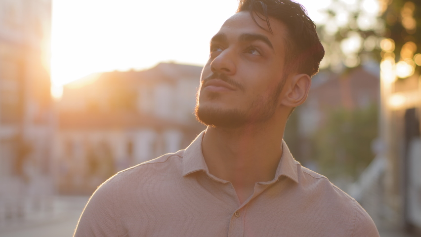 Close-up ethnic indian male happy inspired bearded face. Portrait arabic hispanic business man young adult pensive guy stands in city on street background of sunset sun dreaming thinking enjoying rays