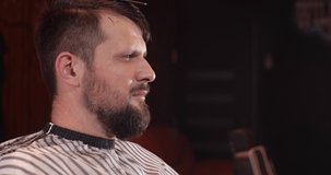 Close-up of a handsome Caucasian guy sitting in a hairdressing salon. An unrecognizable barber shaves a bearded man's head with a trimmer. 4K 50 fps slow motion