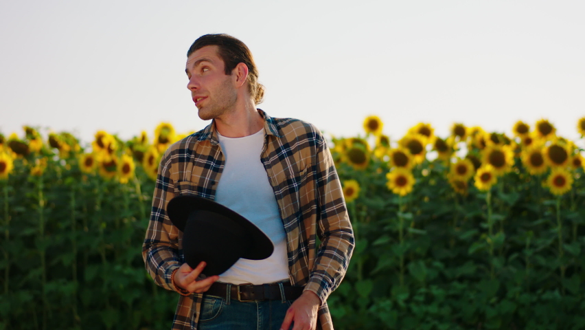 A very attractive looking man with brown hair swearing a checkered shirt and a cowboy hat is smiling while being in a beautiful sunflower field. 4k