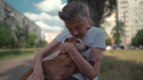 Senior 90-year-old woman with gray hair and deep wrinkles sits outdoors In assisted living facility on bench with small dachshund dog. Old female hugs and cuddles pet in the park on a bench