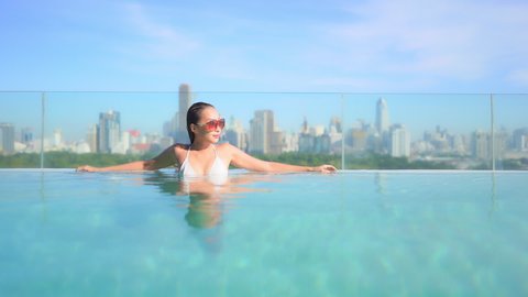 Sexy lady leaning on the edge of a rooftop infinity pool, a beautiful young woman enjoys the sun and water with Bangkok city skyline behind her, Thailand urban vacation