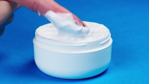Female hand taking moisturizing foam cream for face or body on blue background. Bowl with soft cream close-up, cosmetic texture. Beauty and health, skin care concept.