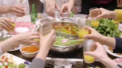 Several people toasting to celebrate, eating Yin-Yang hotpot, vegetables, meatballs, gimbap, beer, drinks, etc. around a round table