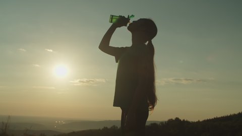 Silhouette of slim active sporty fit asian woman with ponytail drinking water from bottle, refreshing herself after successful intense workout while resting on mountain top in rays of warm setting sun