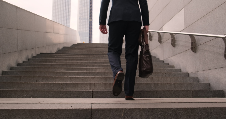 Back view Businessmen put on a black suit at the office. The man holding business handbag with office formal suit walking outdoor in urban city. Legs of a business man wearing brown shoes walking up
