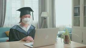 Excited Asian Woman Wearing Protection Face Mask And Holding A University Certificate, Smiling To Camera. Pretty Female Graduate Wearing A Graduation Gown And Cap Sitting On The Living Room

