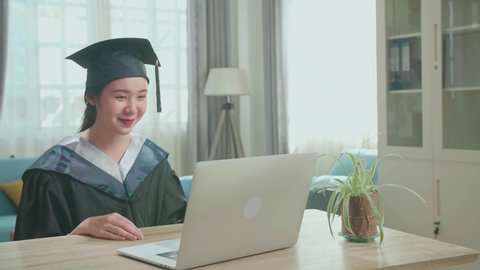 Excited Asian Woman Thumb Up And Showing Off A University Certificate To The Family During An Online Video Call. Pretty Female Graduate Wearing A Graduation Gown And Cap Sitting On The Living Room
