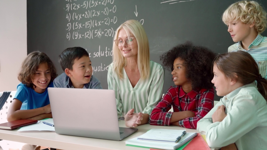 Happy diverse junior school children students gather at teacher table teaching kids looking at laptop computer using online software learning web education tech study together at class in classroom. Royalty-Free Stock Footage #1079702549