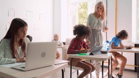 Happy smiling Caucasian teacher explaining encouraging Afro American schoolgirl using tablet device sitting in classroom with group of diverse schoolchildren. Modern education technologies concept. - Powered by Shutterstock - Get 15% off with code: PIKWIZARD15