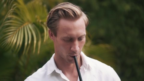 Portrait of young beautiful man smoking in slow motion. Close up video of pretty face Caucasian man smoking hookah outdoor, relaxing after hard day. Art of hookah smoking. High quality FullHD footage