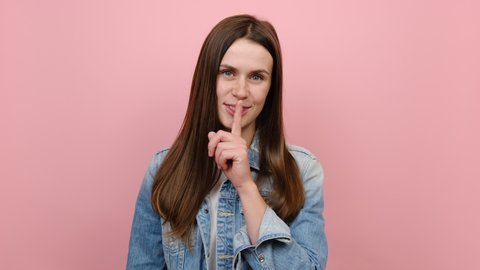 Secret young millennial female 20s years old look aside say hush be quiet with finger on lips shhh gesture close her mouth lock, wears denim jacket, isolated on pastel pink color background studio