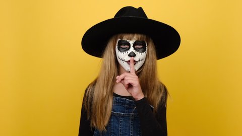 Portrait of secret fun girl kid with Halloween scary makeup mask wears black hat, say hush be quiet with finger on lips shhh gesture, posing isolated over yellow color background wall in studio