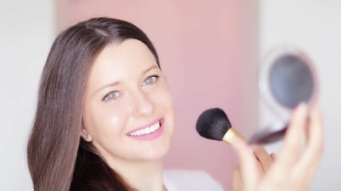 Woman with makeup brush and compact mirror applying cosmetic powder and smiling, face portrait of beautiful model on pink background, natural make-up idea, cosmetics and skincare product commercial.
