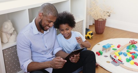 Mixed-race boy spend time with dad family sit on floor in playroom using digital tablet, having fun on internet, enjoy new cool amusing mobile application, play video games online. Modern tech concept