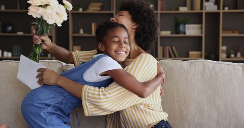 African female sit on sofa at home receive congratulations on Mothers Day, get flowers and postcard from little loving daughter. International Women Day, live events and birthday celebrations concept