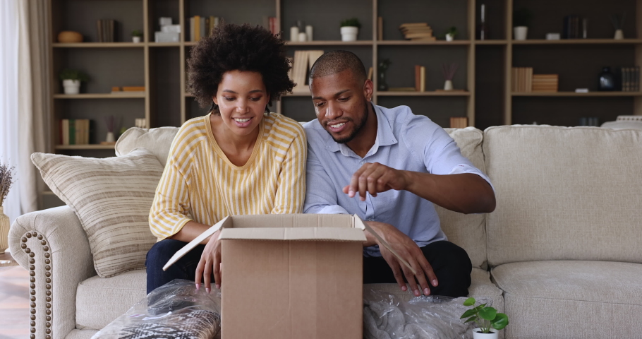African couple sit on sofa at home unpack parcel box, take out bought online fashion winter clothes. Happy satisfied clients of e-commerce retails services. Shoppers family, remote e-shopping concept Royalty-Free Stock Footage #1079720255
